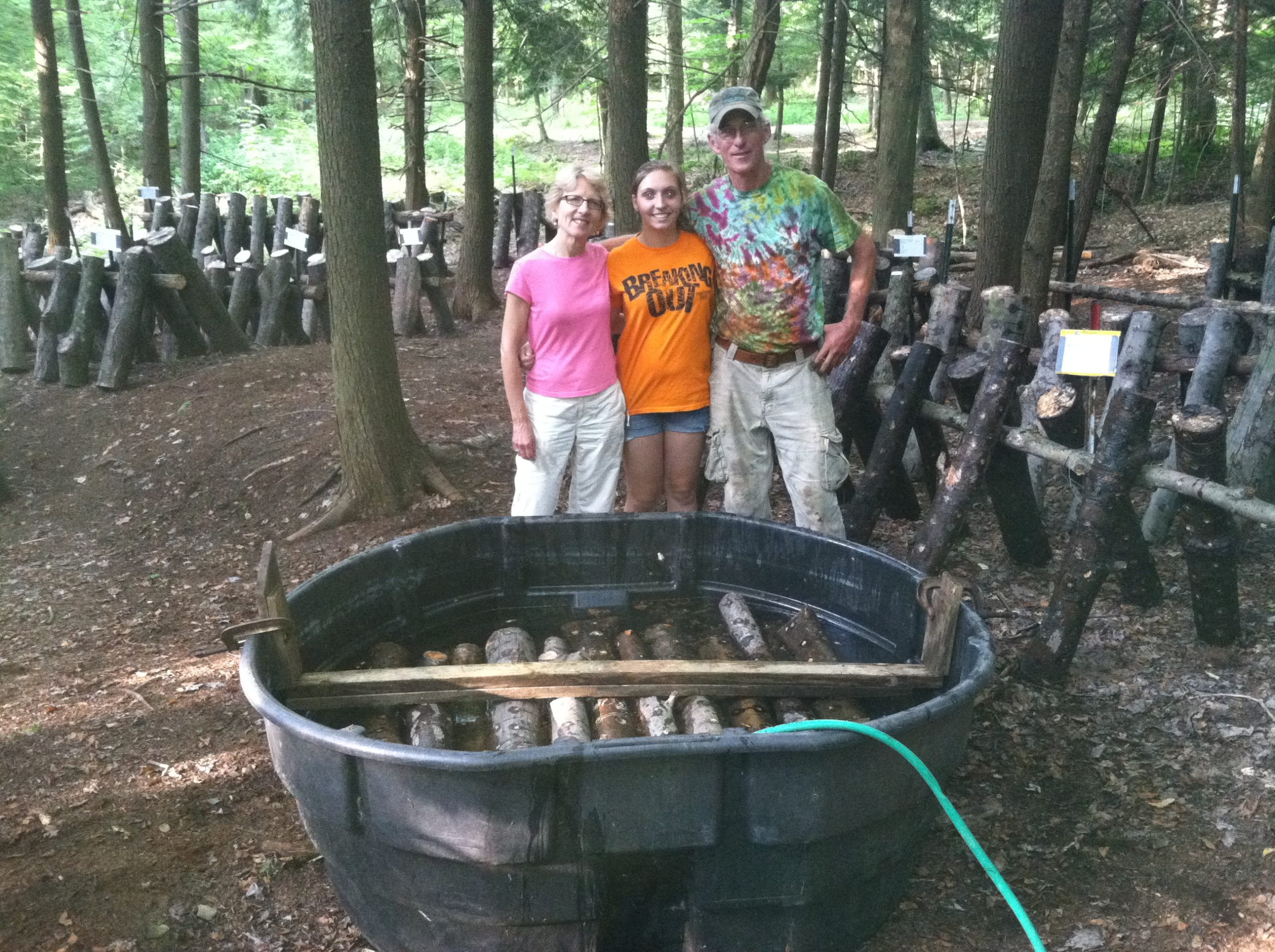 Three people stand with a large tub in front of them, filled with water and soaking logs.