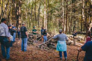 A group of people stands in a forest clearing, listening to a speaker near a pile of logs used for shiitake cultivation. 
