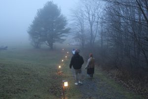 Three people walk along a foggy path lined with small lanterns. The immense pine tree on the left contrasts with bare trees on the right, leading to a house in the background. The scene invites pause, enveloped in misty serenity.