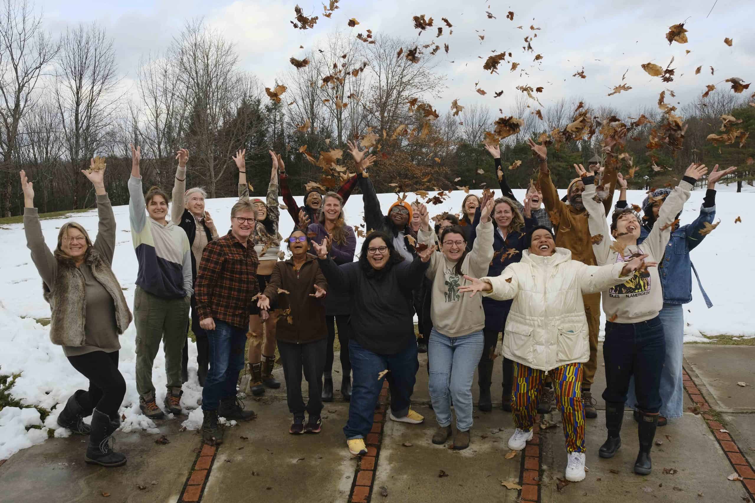 A joyful community gathers outdoors, tossing leaves in the air. Against a backdrop of snowy ground and barren trees, everyone is dressed in winter clothing, smiling and celebrating the moment together.