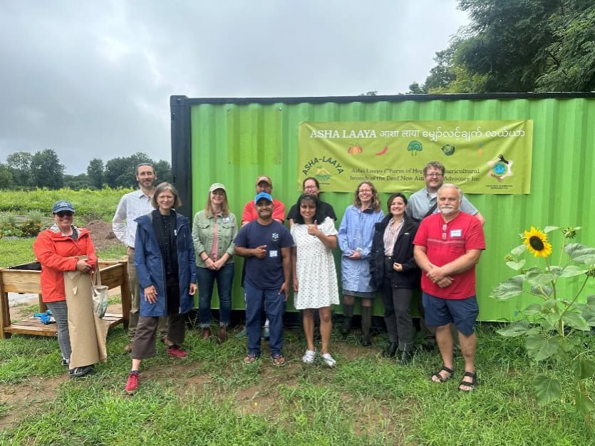 A group of twelve people stand smiling in front of a green container with a yellow banner that reads ASHA LAAYA in both English and another language. They are outdoors on a cloudy day, and a sunflower is visible beside them.