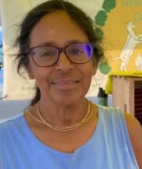 A person with glasses and a double necklace, wearing a blue top, smiles at the camera, embodying the spirit of Farmer Wellness. In the background, a colorful sign with illustrations captures the essence of springs vibrant renewal.
