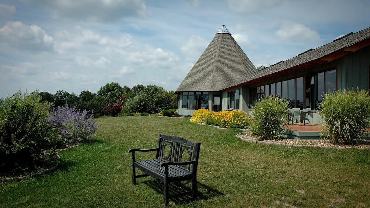 A scenic view of a unique house with a conical roof surrounded by lush greenery and colorful flowers. Theres a wooden bench on the lawn, and the sky is partly cloudy. The house has large windows and a charming, rustic appearance.