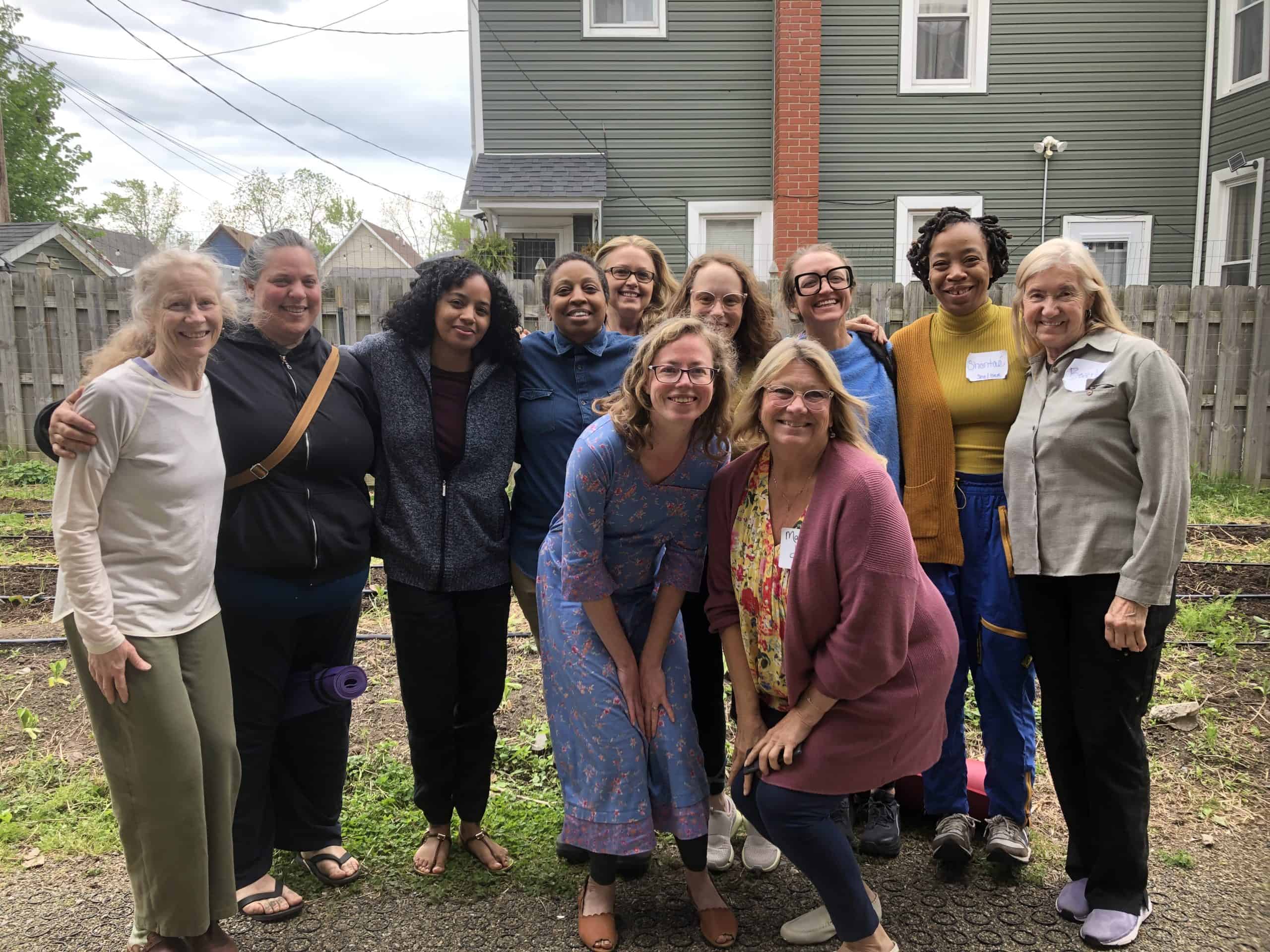 A group of people smiling and posing together outdoors in front of a house, seemingly enjoying a Community Lunch. Some are standing, while two are crouching at the center. They’re dressed casually for this gathering. The background displays a wooden fence and lush greenery.