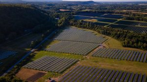 Aerial view of large solar panel fields utilizing agrivoltaics, surrounded by trees and farmland, with hills and a distant horizon under a clear sky.