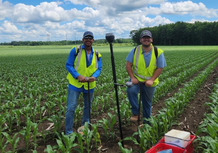 Two farmers in yellow safety vests, one wearing a blue shirt and the other a gray shirt, stand in a lush green cornfield under a partly cloudy sky. They are holding tools related to manure management and surrounded by equipment. Dense greenery and trees are in the background.