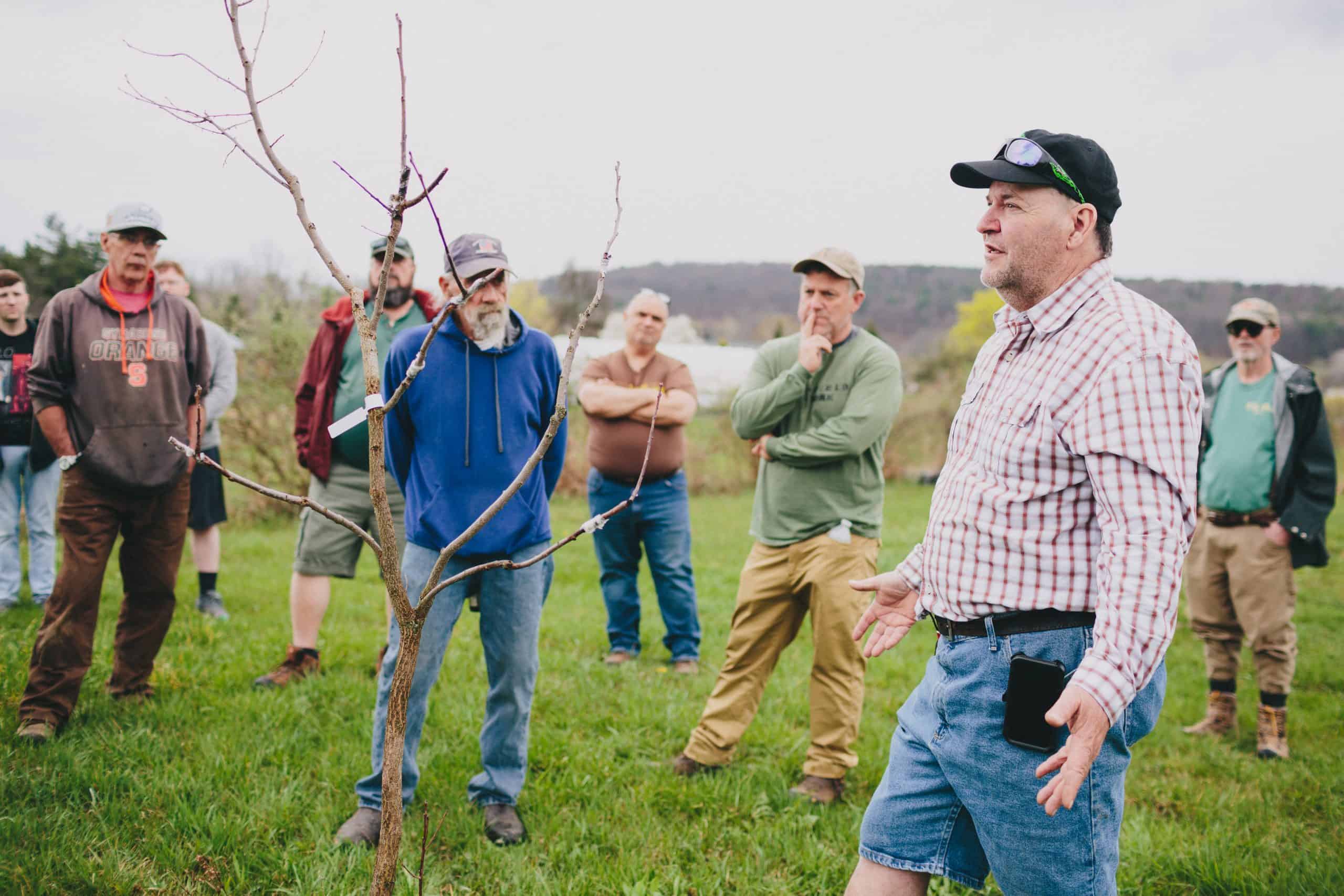 A man in a plaid shirt and shorts leads an Orchard tour at Ort Family Farm, speaking to a group outdoors by a young tree. The group listens attentively, standing on green grass with trees and hills in the background.