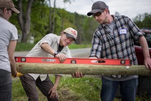 Three people work together outdoors at a farm event to position a wooden pole for livestock fencing, using a red level to ensure it is straight. Trees and a road are visible in the background, and one person wears a nametag that says Jeremy.