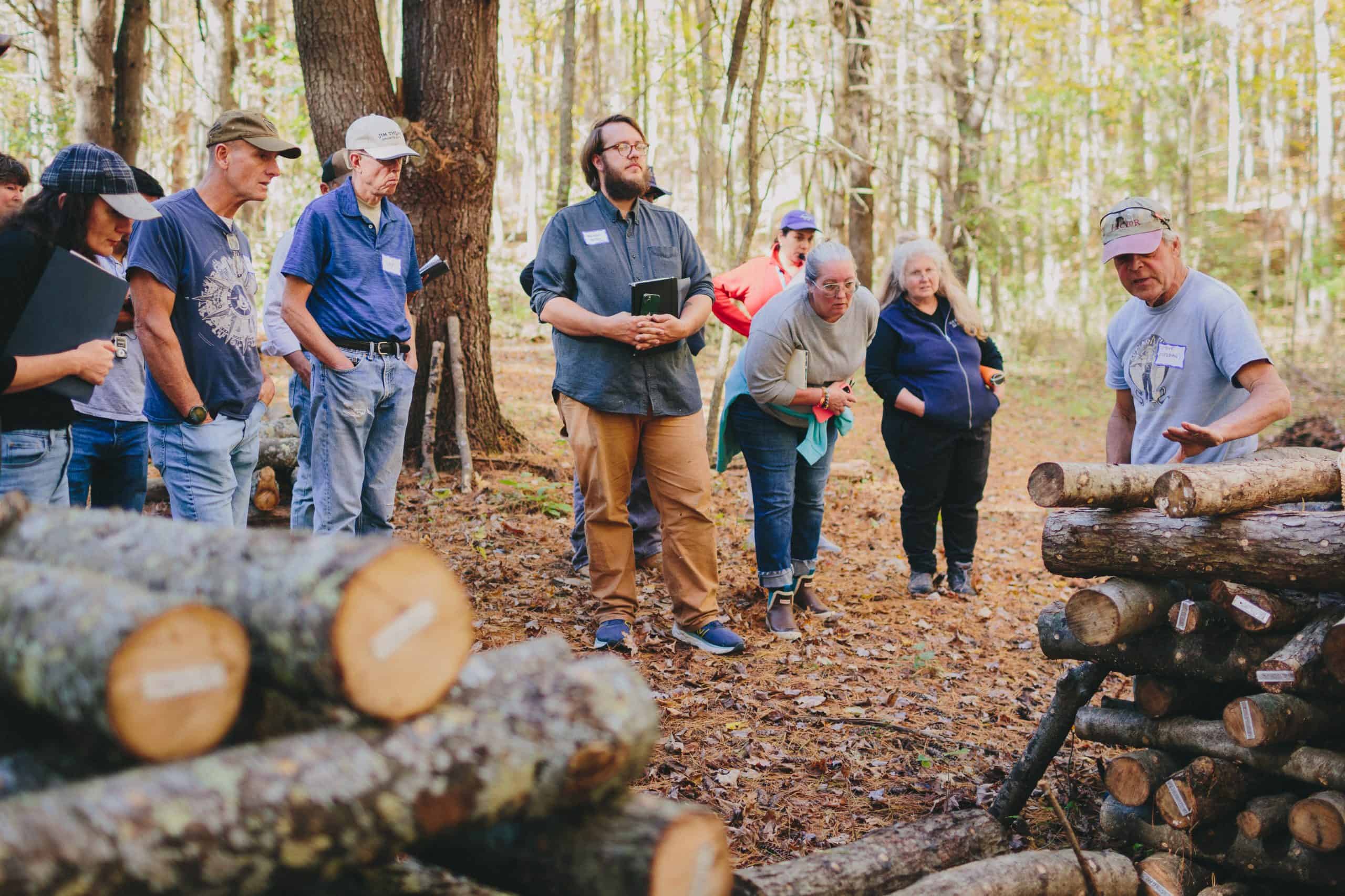 Shiitake Mushroom Cultivation Workshop 1 A group of people stand in a wooded area, attentively listening to a man in a cap leading a workshop on Shiitake mushroom cultivation as he explains something about stacked logs among the sunlit trees.