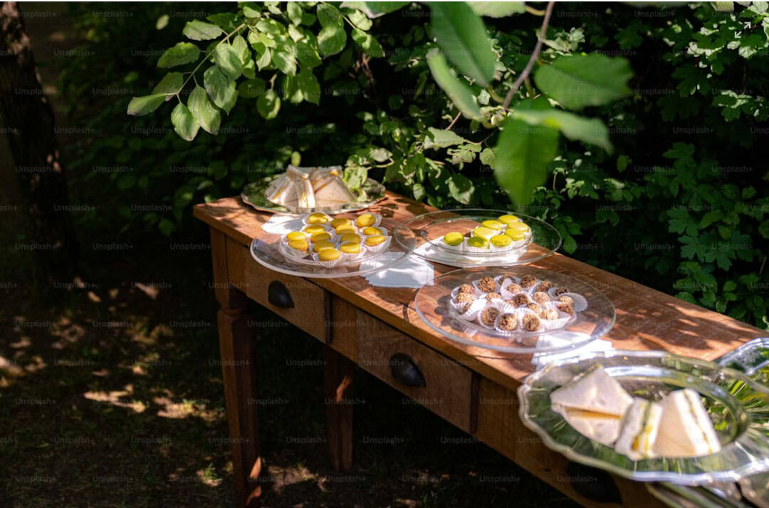 A wooden table outdoors under leafy trees holds glass plates with lemon tarts, sandwiches, and round desserts, arranged for a community farm supper or picnic. Sunlight filters through the leaves above.