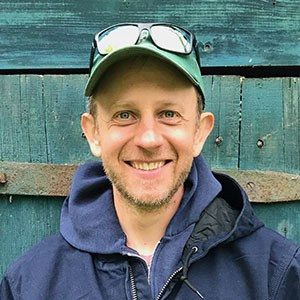 A man wearing a green cap, sunglasses on top of his head, and a blue hoodie smiles while standing in front of a weathered teal wooden wall, possibly after attending one of the small farms courses nearby.