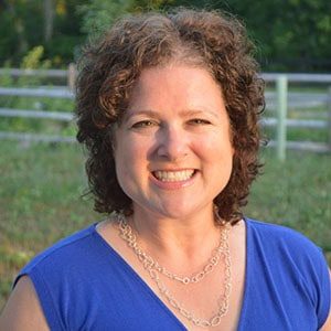 A woman with curly brown hair, wearing a blue top and silver chain necklace, smiles outdoors after attending small farms courses, standing in front of a blurred background with greenery and a wooden fence.