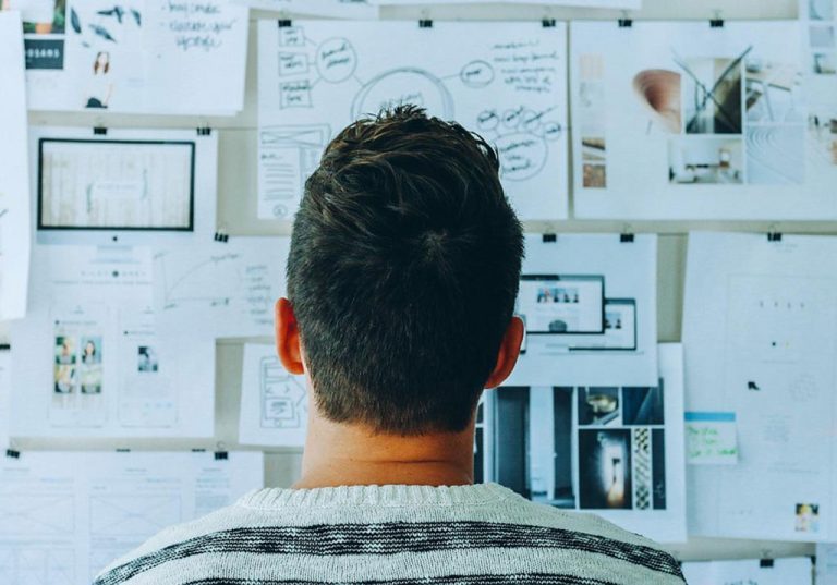 A person with short dark hair in a striped sweater looks at a wall covered with sketches, diagrams, and printed documents. The materials are arranged in a somewhat random manner, suggesting brainstorming or planning in progress, as though they're starting at square one on BF 101.