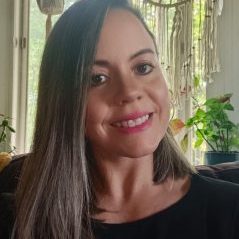A woman with straight, shoulder-length brown hair and a black top smiles at the camera. Behind her are green plants, large windows, and a decorative macrame wall hanging—evoking a cozy, tim shenk-inspired ambiance.